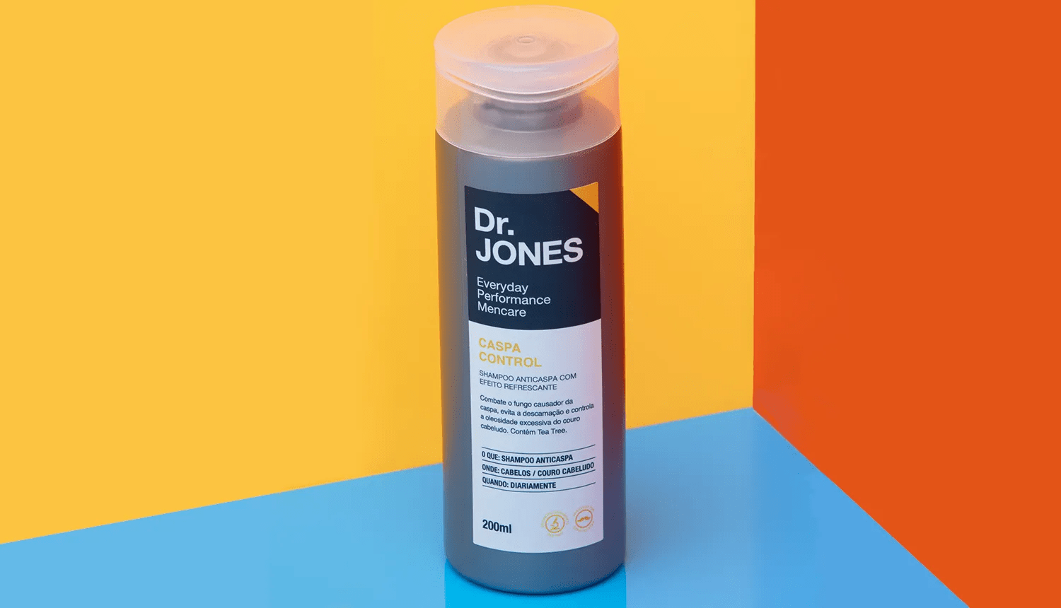 Por dentro do produto: Climbazol - Dr. JONES. Em fundo geométrico azul-claro, laranja e dourado, foto em destaque da embalagem do shampoo anticaspa Caspa Control, da Dr. JONES.
